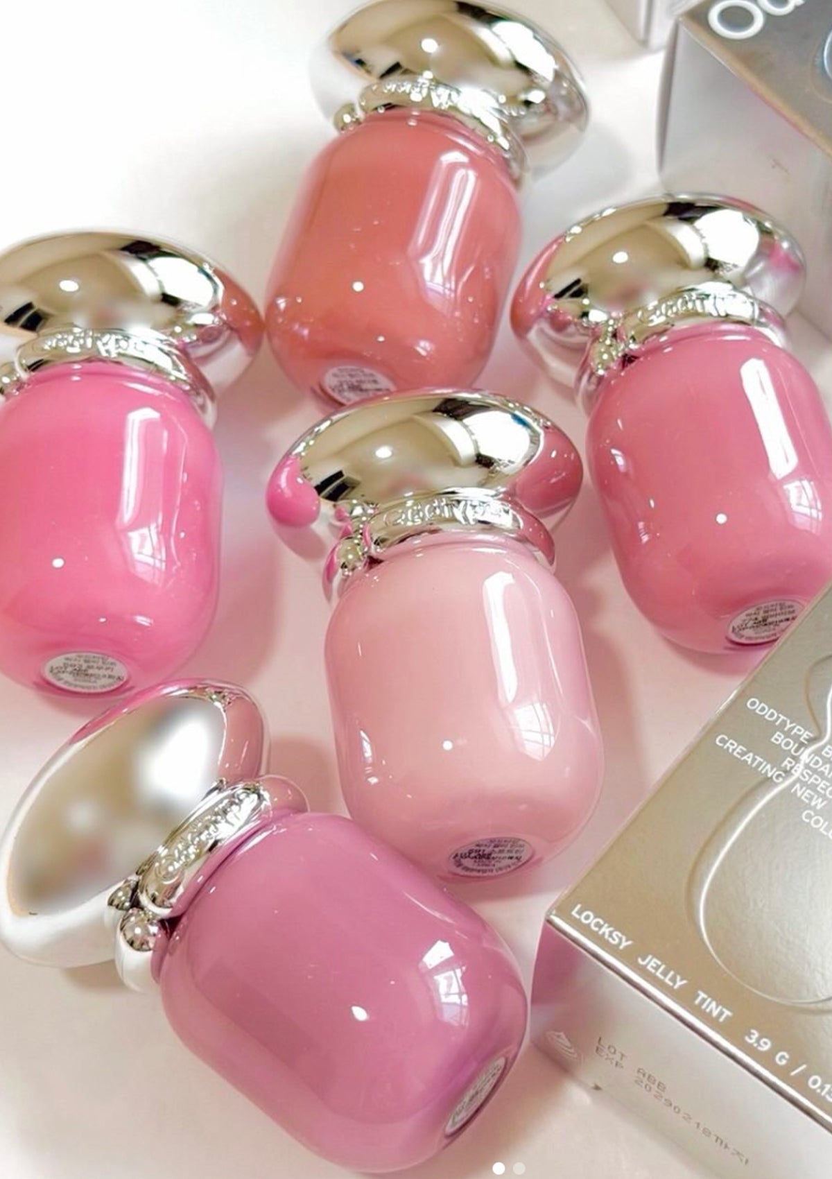 Six bottles of glossy pink nail polish in various shades, arranged on a light surface with a box partially visible in the corner.
